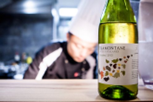 A bottle of white wine on a wooden counter, a blurred cook in a kitchen behind it.